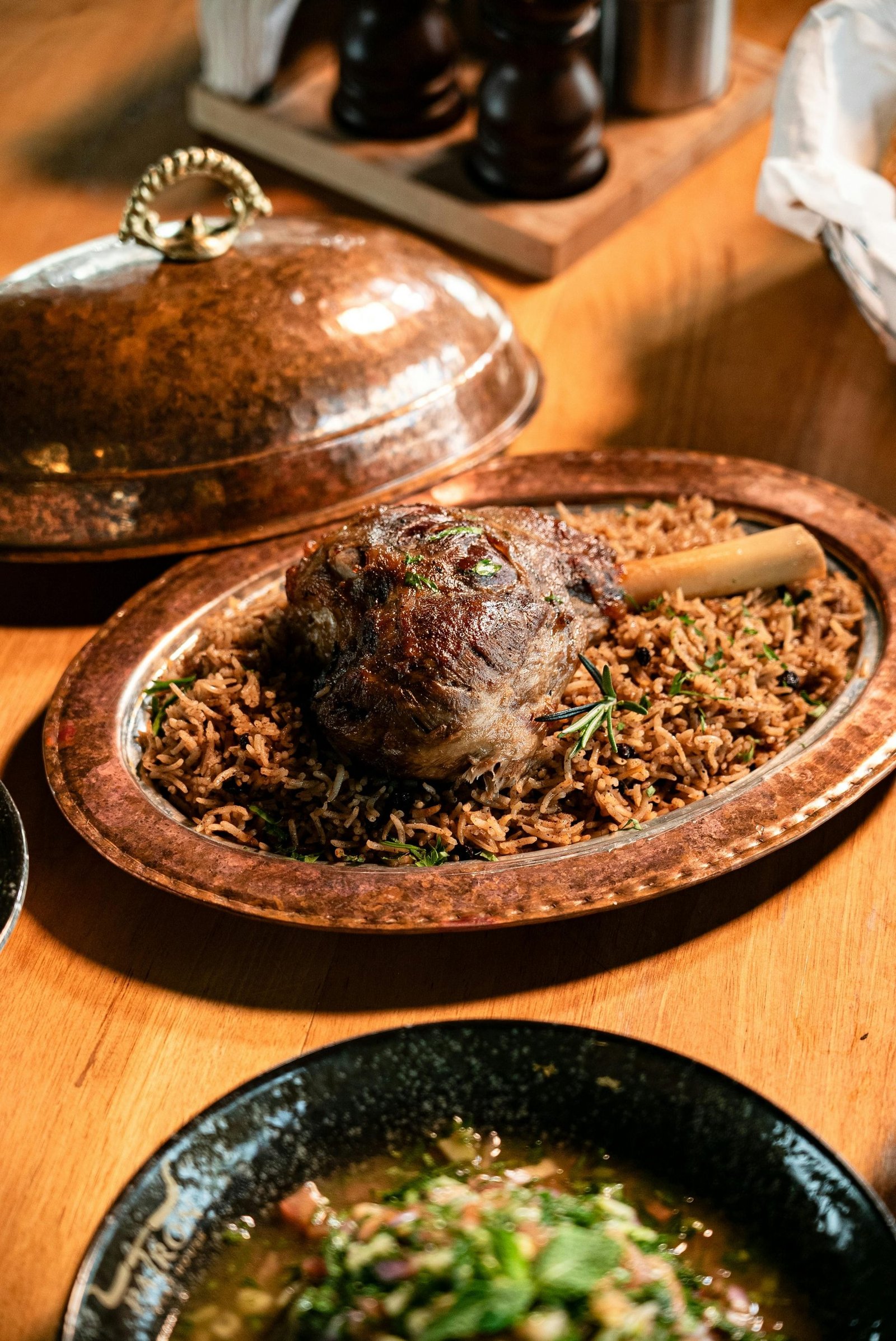 Whole roasted lamb shank on a copper platter with seasoned rice, fresh rosemary, and an Arabic copper dome lid
